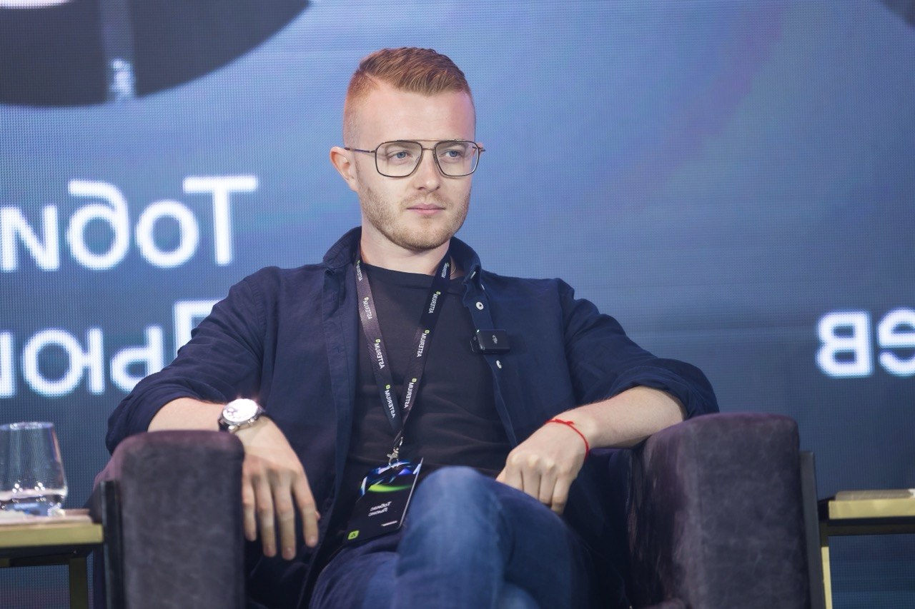 Tobias Jack Lewis, in glasses and a dark shirt with a conference lanyard, sits in an armchair on a panel stage